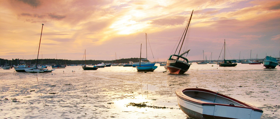 Small boats resting on the mudflats at low tide in Poole Harbour during a vibrant sunset, with warm orange and pink light reflecting across the water.