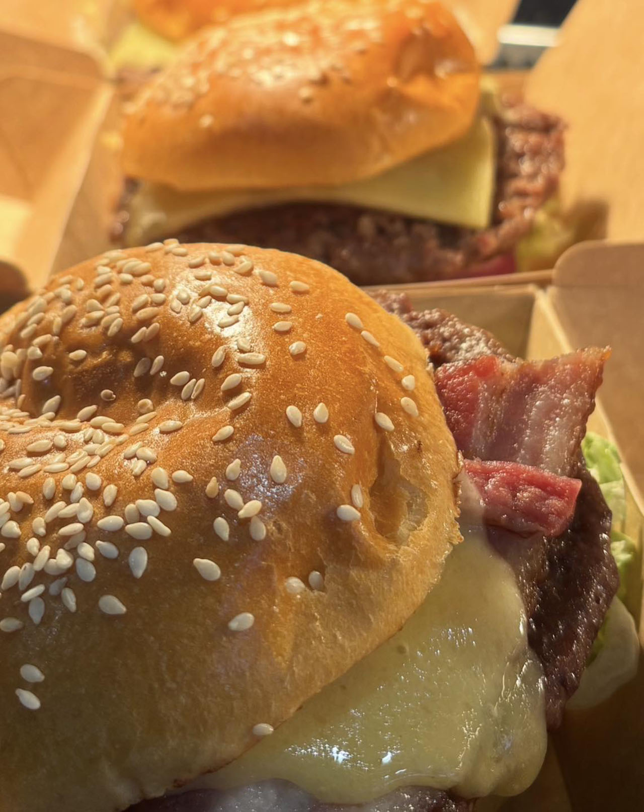 Close-up of two gourmet burgers in cardboard trays, featuring sesame seed buns, melted cheese, and crispy bacon.